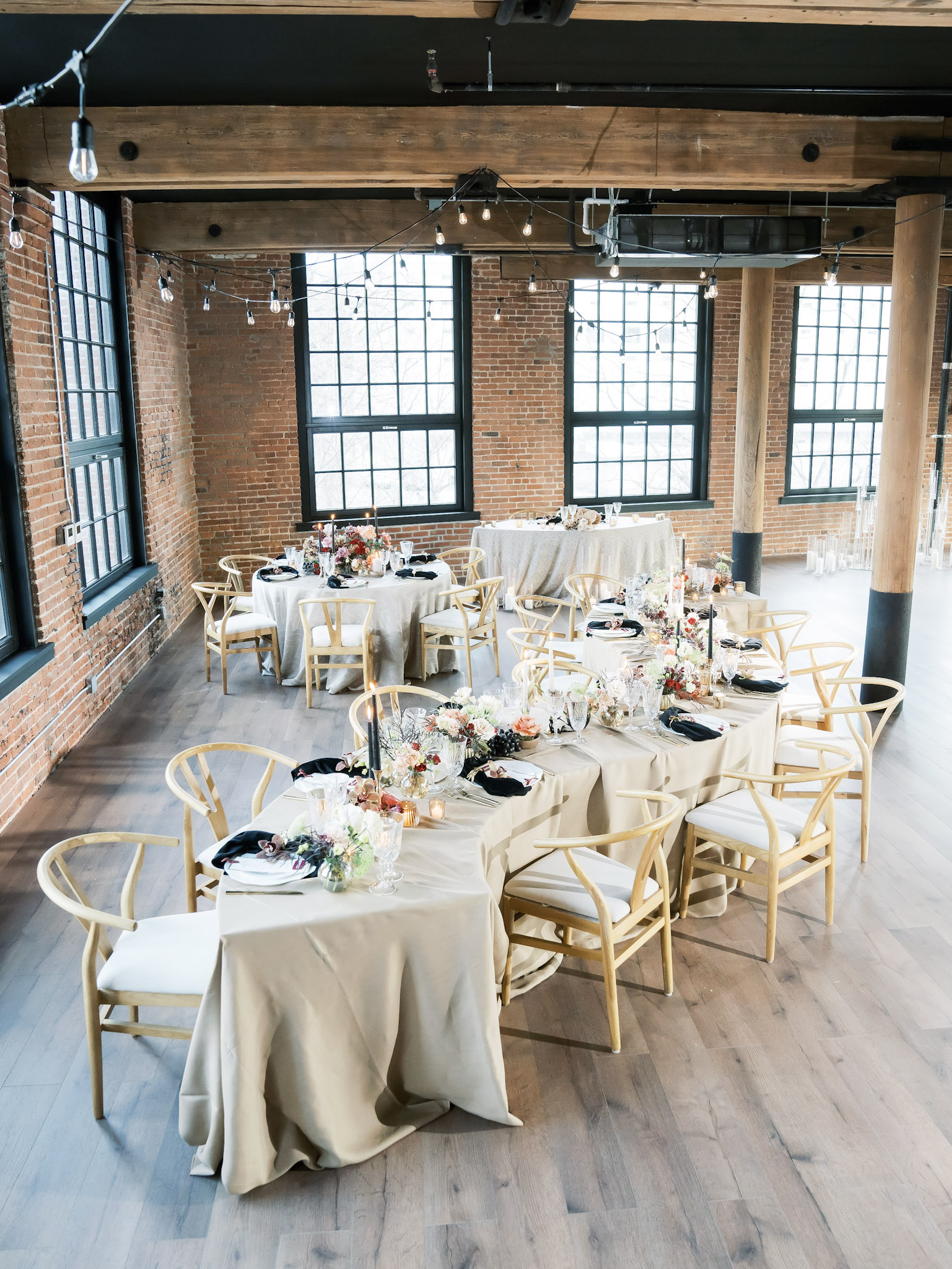 Wedding reception with long tables, wooden chairs, and floral centerpieces by industrial windows